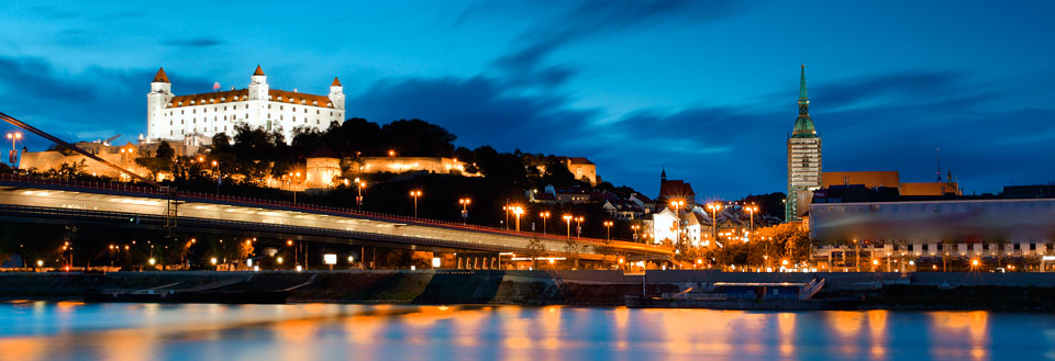 Et aftensbillede af Donau i Bratislava med en bro i forgrunden og et oplyst slot på en bakketop i baggrunden.