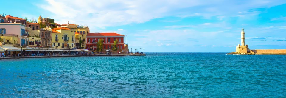 Farverige huse langs en kyst med en fyr i baggrunden, klart blåt vand og skyfri himmel.