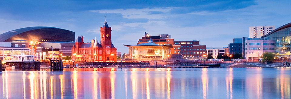 Cardiffs vandfront med moderne bygninger og et gammelt tårn reflekteret i vandet.