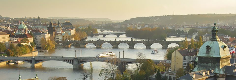 Billede af Prags skyline med Karlsbroen og floden Moldau. Bygninger og broer fremstår i aftensolens lys.