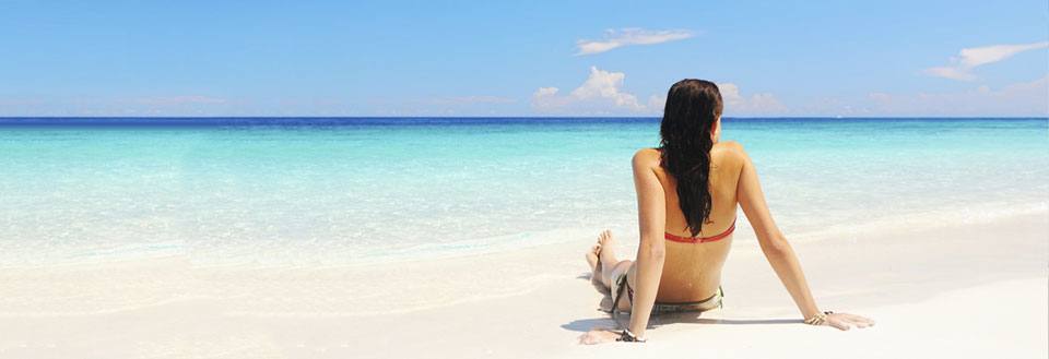 En person sidder på en hvid sandstrand og kigger ud over en azurblå hav.