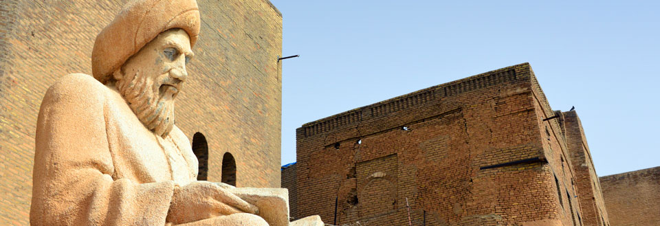 Billedet viser en sandstensstatue af en mand med skæg og turban foran en murstensbygning under en klar blå himmel.