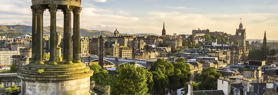 View over Edinburgh med klassiske bygninger, monumenter og grønne områder under en klar himmel.