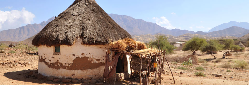 Traditionel hytte med stråtag i en tør landsby med bjerge i baggrunden.