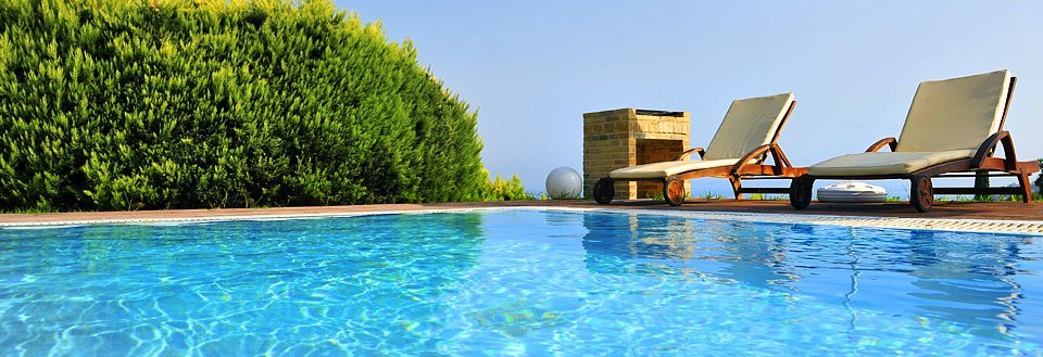 Et fredeligt poolområde med to solsenge, klart blåt vand og en frodig grøn hæk i baggrunden.