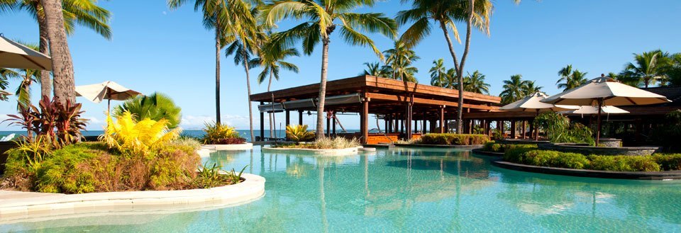 Luksuriøst feriested med svømmebassin omgivet af palmetræer og liggestole under parasoller.