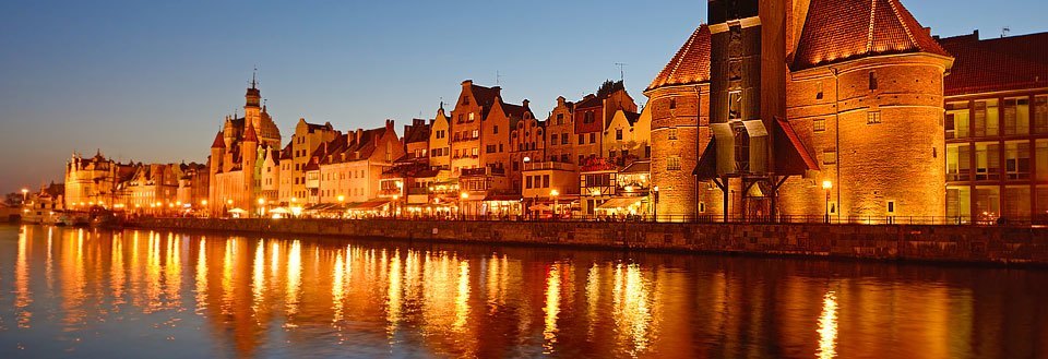 Aftenbillede af Gdansk ved floden, lys fra bygninger spejler sig i vandet.
