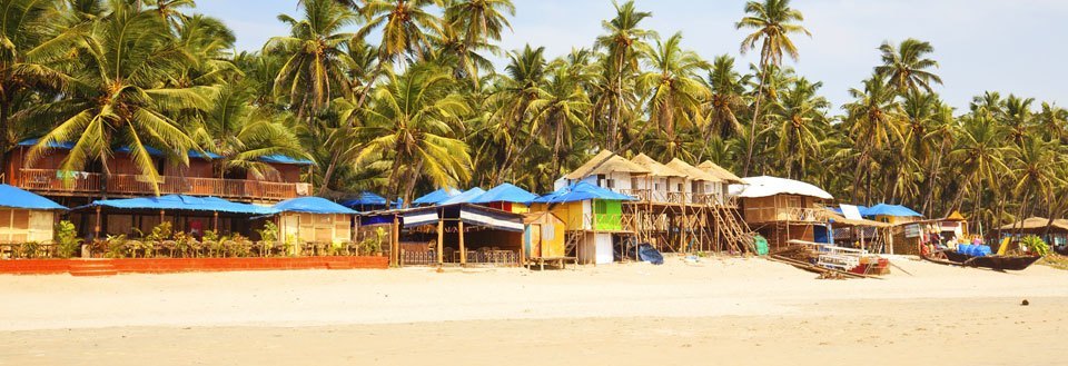 En tropisk strand i Goa med kokospalmer i baggrunden og små hytter foran. Nogle både ligger på sandet.
