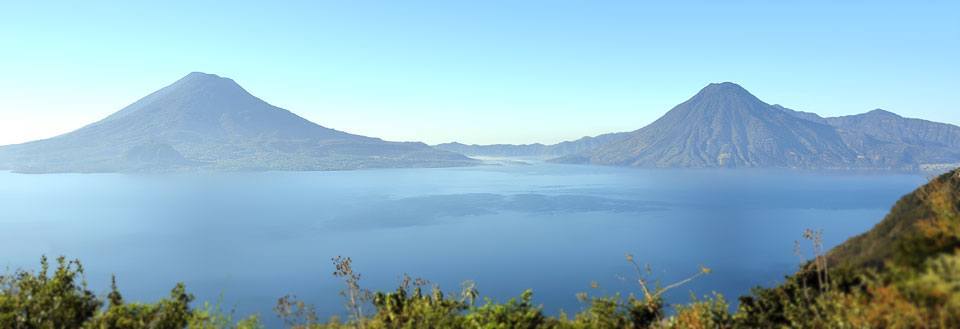 Panoramaudsigt over en rolig sø med imponerende vulkaner i baggrunden og en klar blå himmel.
