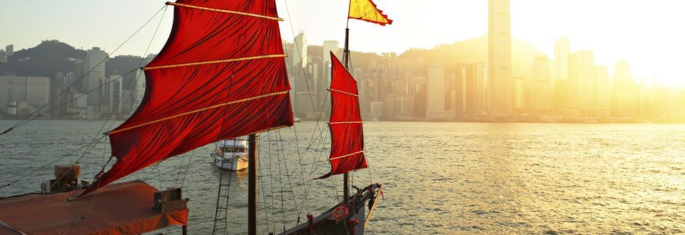 En traditionel sejlbåd med røde sejl flyder foran Hong Kongs byskyline i solnedgangens gyldne lys.