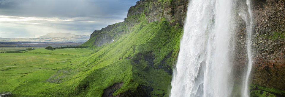 Et frodigt landskab i Island, med et kraftigt vandfald på en grønklædt klippevæg under en himmel med let skydække.
