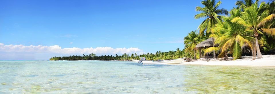 Tropisk strand med palmer, klart vand og en lille båd. Blå himmel med spredte skyer.