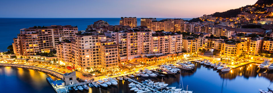 Aftenbillede af Monacos havnefront med oplyste bygninger og lystbåde i marinaen.