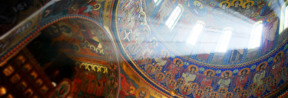 Indvendig kuppel af en kirke med farverige freskomalerier og ikoner, oplyst af naturligt lys fra vinduerne.
