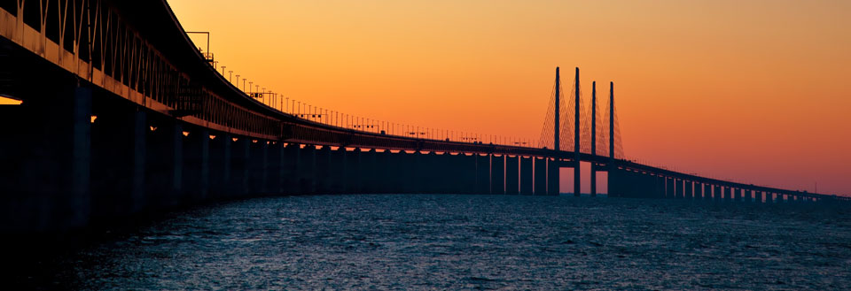 Lang bro over vand ved solnedgang med orange himmel.