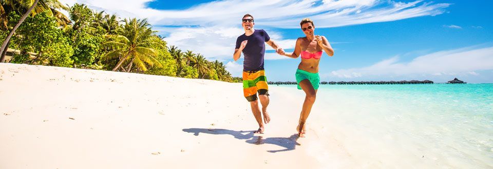 To personer løber smilende på solrig strand med hvidt sand og palmetræer i baggrunden.