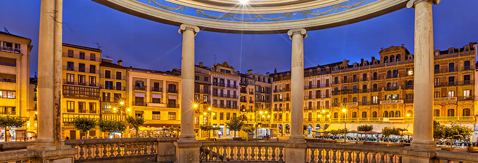 Et stemningsfuldt aftenbillede af en plads i Pamplona  omkranset af klassiske bygninger belyst af gadebelysning.