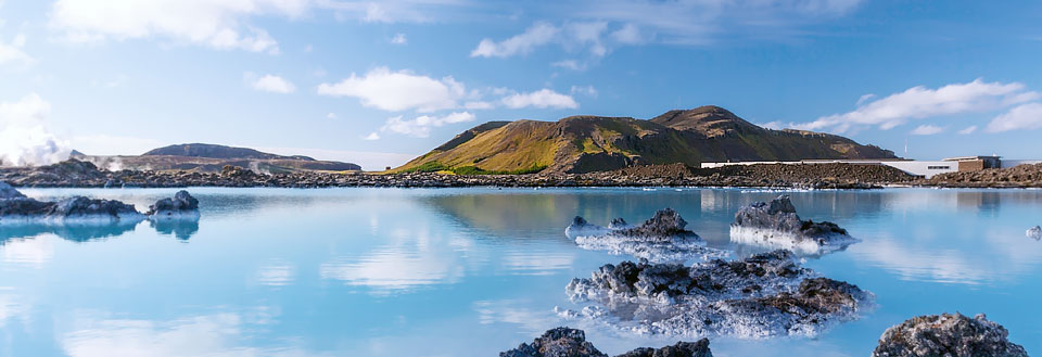 Et storslået landskab med et varmt, turkisblåt geotermisk vandbassin omgivet af vulkanske klipper og grønne bakker.
