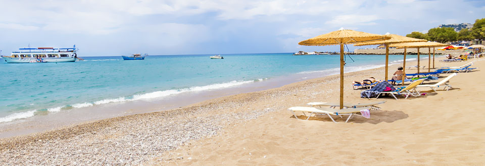 En solrig strand med parasoller, liggestole og et par personer. Klar blå himmel og smukke båd i det fjerne.
