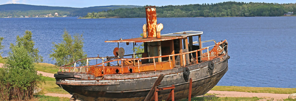 En rusten gammel båd på land med en skov og en sø i baggrunden.