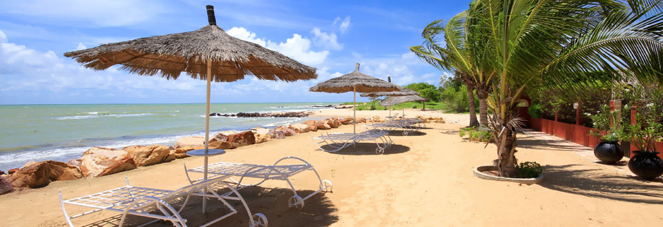 En solrig strandpromenade med parasoller, liggestole, og et azurblåt hav i baggrunden.