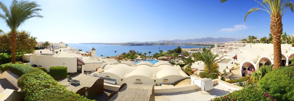 Panoramaudsigt over et ferieresort med hvidkalkede bygninger, palmer, en swimmingpool og havet i baggrunden.