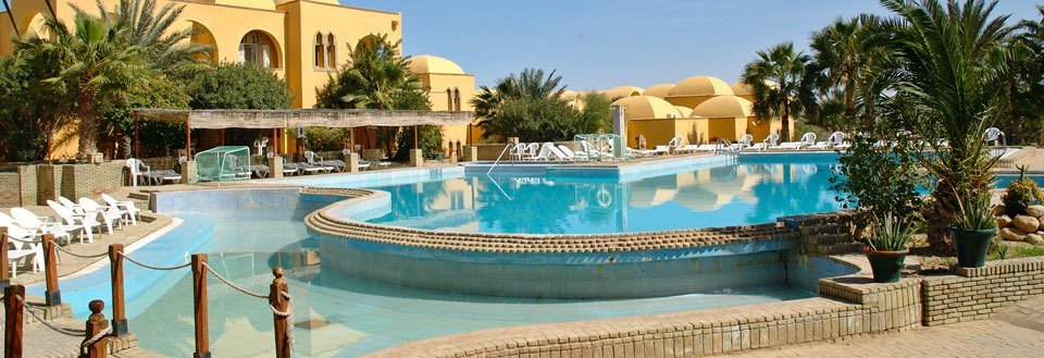 Luksuriøst resort med en stor svømmepøl, omgivet af palmer og hvilestole under solen.