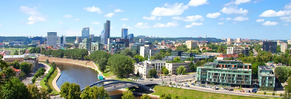 Panoramaudsigt over Vilnius med moderne bygninger, en flod og en bro under en klar blå himmel.