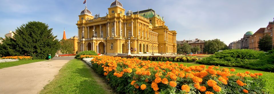 Et storslået gult palads med flag, omgivet af en frodig grøn have og orange blomster under en klar blå himmel.