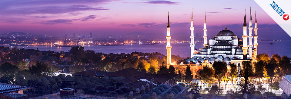 Aftenbillede af Istanbul med en historisk moské med tårne foran en by med lys og vand.