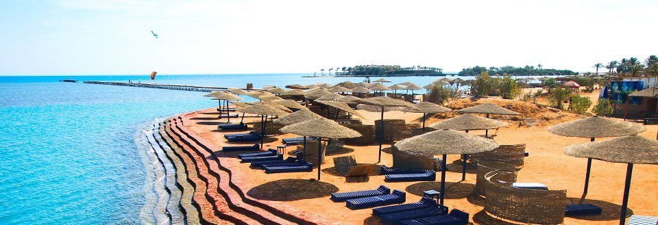 Strand med parasoller og liggestole foran klart blåt vand. Ferieresorts og kitesurfer i det fjerne.