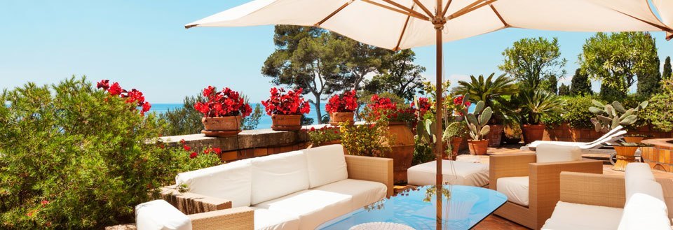 Solrig terrasse med hvide havemøbler, parasol, blomsterkrukker og middelhavstræer.