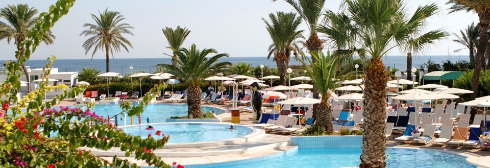 Luksuriøst feriested med swimmingpools, palmer og parasoller ved en solrig kystlinje.