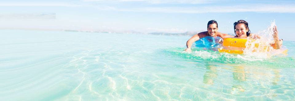 To personer smiler og svømmer med oppustelige baderinge i krystalklart havvand.