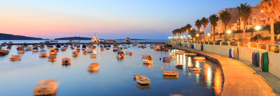 Solnedgang over en havn på Malta med mange små både. Vandet spejler den varme himmel og kystlinjen er belyst.