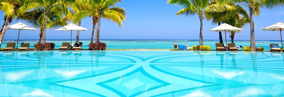 En swimmingpool med udsigt til havet, omgivet af palmetræer og solsenge under parasoller.