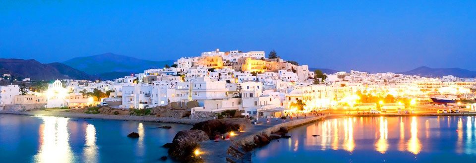 En malerisk kystby på Naxos ved skumring med oplyste hvide bygninger og reflekterende vand.