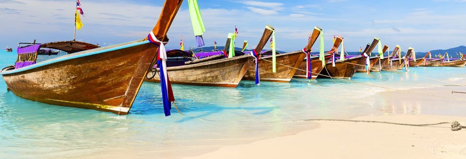 Traditionelle træbåde på Phuket med farverige bånd fortøjet på en tropisk strand med krystalklart vand.