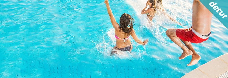 Glade mennesker hopper og leger i en funklende blå swimmingpool på en solrig dag.