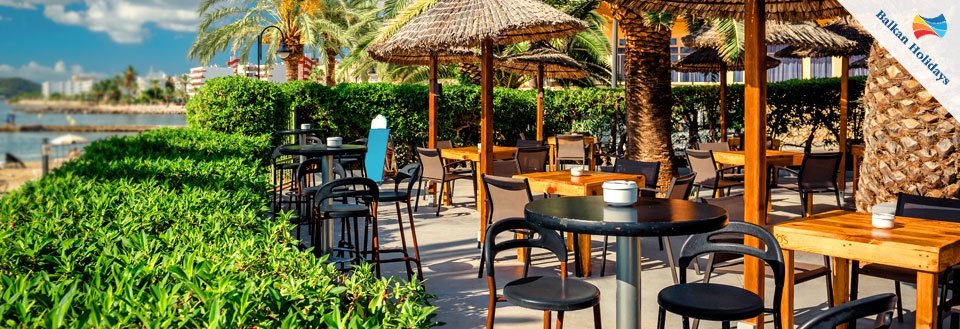 En solbeskinnet udendørs café ved stranden med tomme stole og borde, palme parasoller og grønne buske.