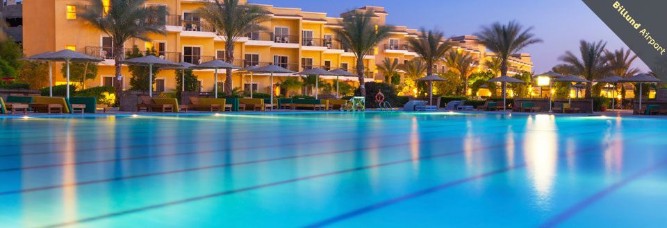 Et luksuriøst hotel og en oplyst swimmingpool der reflekterer aftenhimmelen.