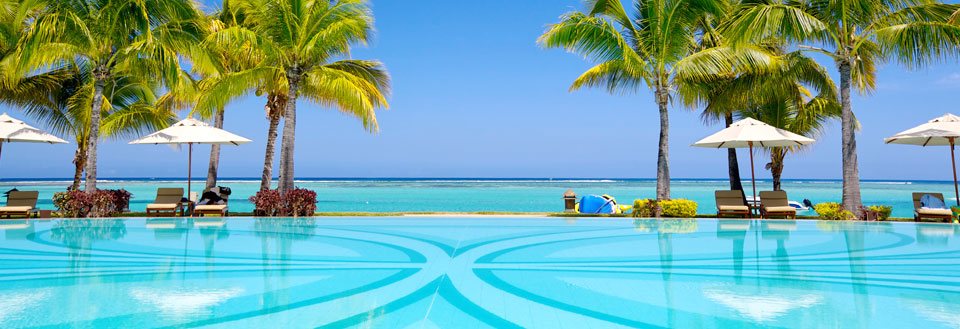 Luksuriøst resort med svømmebassin foran en tropisk strand med palmer og parasoller.