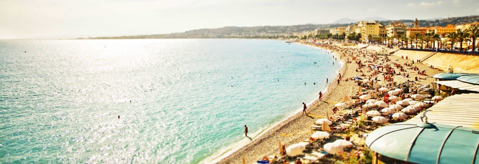 En solrig strandpromenade med mange mennesker og parasoller langs det azurblå hav.