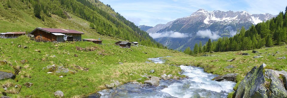 En idyllisk bjergdal med træhytter, en strømmende flod og grønne enge mod en baggrund af sneklædte bjerge.