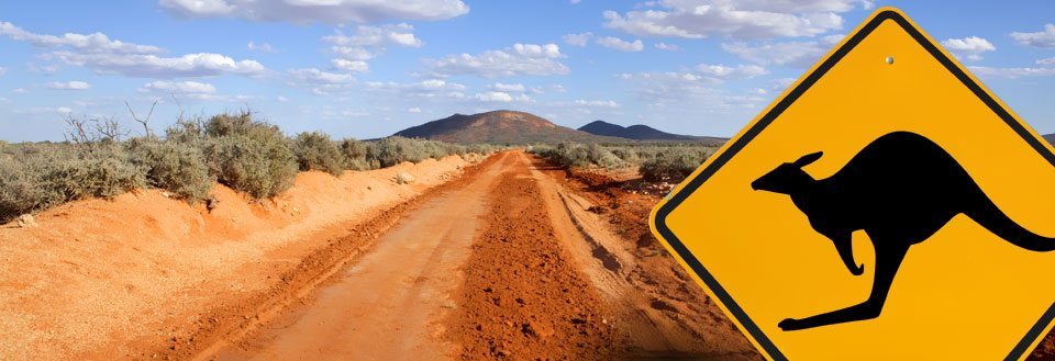 Rød jordvej der strækker sig mod et bjerg under en blå himmel, med skilt for kænguru.
