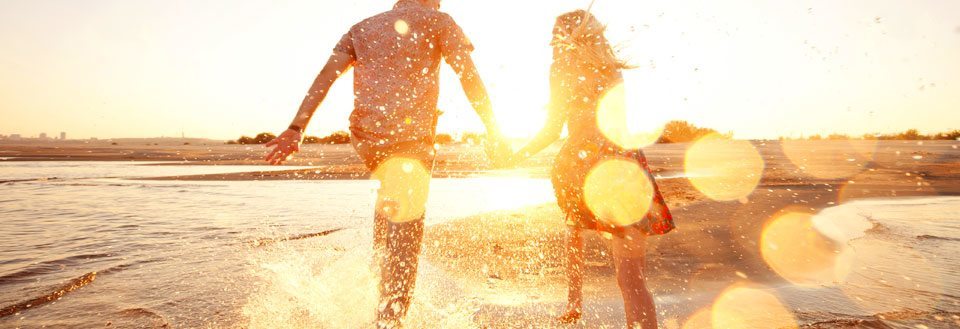 To personer holder i hånden og løber gennem vandkanten på stranden ved solnedgang.