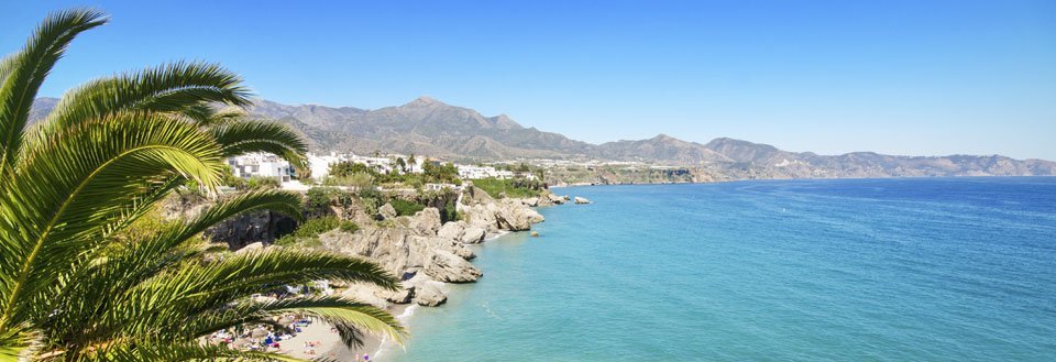 Panoramaudsigt over en klippefyldt kystlinje med klart blåt hav, palmetræer og hvidkalkede bygninger mod bjerge i baggrunden.