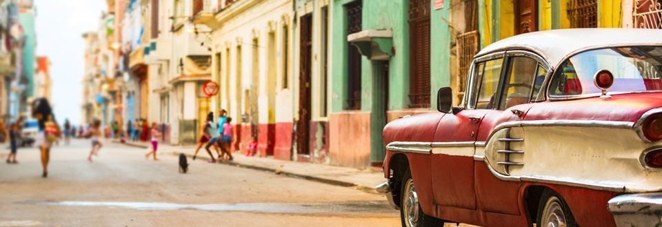En farverig gade med gamle bygninger og en klassisk bil foran. Mennesker gå og snakke langs fortovene.