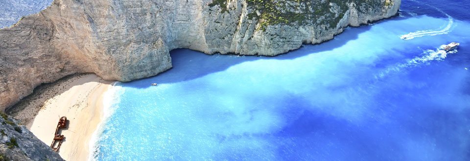 Storslået strand med høje klipper og krystalklart blåt vand. En båd sejler i nærheden.