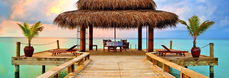 En træbrygge fører ud til en overdækket lounge med stråtag ved et turkis hav under en farverig himmel.